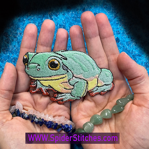 Könnte beinhalten: Grüne und gelbe gestickte Frosch-Aufnäher mit roten Akzenten. Der Aufnäher liegt in der Handfläche.