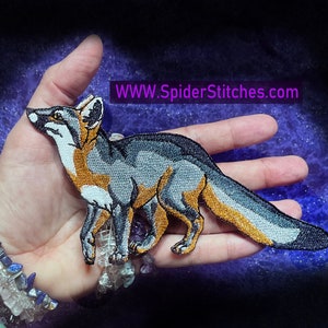 Könnte beinhalten: Ein gestickter Aufnäher eines Fuchses in Grau und Orange mit einer weißen Brust und einer schwarzen Nase. Der Aufnäher befindet sich auf einer Hand mit einem Armband aus bunten Steinen. Der Text "WWW.SpiderStitches.com" befindet sich in der oberen rechten Ecke des Bildes.