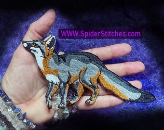 May include: An embroidered patch of a fox, featuring grey and orange fur, a white chest and a black nose. The patch is displayed on a hand wearing a bracelet of colourful stones. The text "WWW.SpiderStitches.com" is in the top right corner of the image.