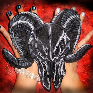 Puede incluir: Un parche bordado en blanco y negro de un cráneo de carnero con grandes cuernos. El parche está sostenido en las manos de una persona con esmalte de uñas azul.