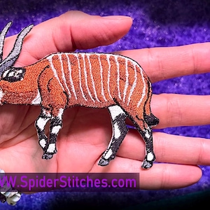 Può includere: Una toppa ricamata con un bongo, una specie di antilope, marrone e bianco, con corna lunghe. La toppa è su una mano con un braccialetto al polso. Il testo "www.spiderstitches.com" è sotto la toppa.