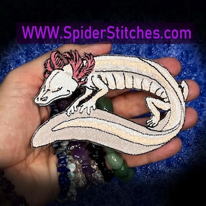 May include: An embroidered patch featuring a cream-colored axolotl with pink gills and black outline details. The patch is shaped like a curled axolotl and includes the text WWW.SpiderStitches.com printed in purple at the top of the image.