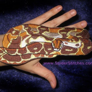 Puede incluir: Un parche bordado de una serpiente enroscada con la barriga blanca. La serpiente tiene un diseño de patrón marrón y amarillo. El parche está en una mano.