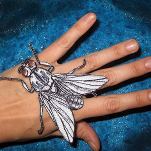 Könnte beinhalten: Ein gestickter Fliegen-Patch in Weiß und Grau mit braunen Augen. Der Patch befindet sich auf der Hand einer Person.