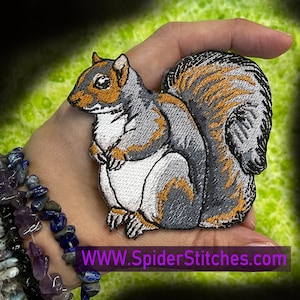 Puede incluir: Parche de hierro bordado de una ardilla gris con vientre blanco y detalles naranjas. La ardilla está sentada con la cola enroscada detrás de ella. El parche está en una mano, y el texto "WWW.SpiderStitches.com" es visible debajo del parche.