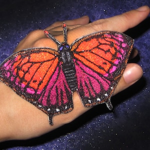 Peut inclure: Un écusson brodé en forme de papillon rose et orange avec des détails noirs. L'écusson est sur une main.