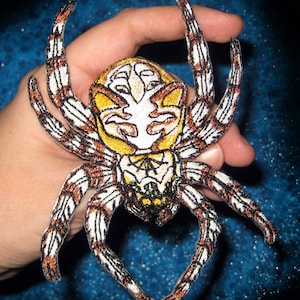 Könnte beinhalten: Ein gestickter Spinnen-Patch in Braun und Weiß mit einem gelben und weißen Gesicht. Die Spinne hat acht Beine und ist erhöht.