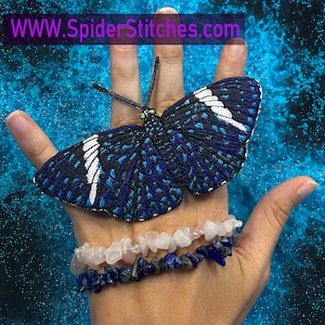 May include: A blue and white butterfly embroidered patch with a black outline. The patch is on a hand wearing two bracelets, one with pink stones and one with blue stones. The background is a blue and white marbled pattern. The text "WWW.SpiderStitches.com" is visible at the top of the image.