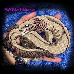 May include: Embroidered patch featuring a stylized, tan sea creature with brown and pink accents. The creature has blue eyes and an open mouth with white teeth. The patch is held in a hand against a blue background. The website www.SpiderStitches.com is visible.