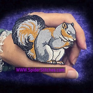 May include: A gray and orange embroidered iron-on patch of a squirrel with its paws clasped together in a praying position. The patch is on a hand with a bracelet.