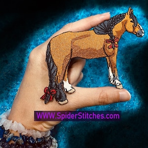 Puede incluir: Un parche bordado que representa un caballo marrón y negro con un lazo rojo. El parche está sostenido en una mano que lleva una pulsera blanca y azul. El texto "WWW.SpiderStitches.com" es visible en la esquina inferior derecha de la imagen.