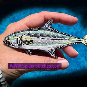 May include: A detailed embroidered patch of a silver and green fish with yellow accents. The fish is depicted swimming to the right. The patch is on a hand, with the text "WWW.SpiderStitches.com" visible below the fish.