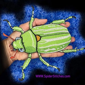 Op de afbeelding: Een geborduurde patch in de vorm van een groene kever met witte strepen, zwarte omlijningen en oranje antennes. De patch heeft een klein oranje accent bij de kop en bevat de tekst WWW.SpiderStitches.com in het paars aan de onderkant.