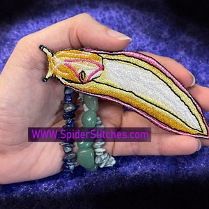 May include: A yellow and pink embroidered patch of a slug with a white underside. The patch is held in a hand with a stack of bracelets on the wrist. The text "WWW.SpiderStitches.com" is visible below the patch.