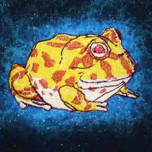 May include: An embroidered frog patch in yellow and orange with red eyes. The frog is sitting on a blue background with white speckles.