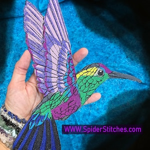 Puede incluir: Un parche bordado de colibrí colorido con detalles en morado, verde, azul y amarillo. El parche presenta un diseño detallado del colibrí en vuelo con las alas extendidas. El texto "WWW.SpiderStitches.com" es visible en la esquina inferior derecha de la imagen.