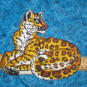 Puede incluir: Parche bordado de un leopardo blanco y marr&oacute;n con ojos azules, acostado sobre un fondo azul brillante.