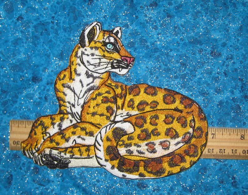 Puede incluir: Parche bordado de un leopardo blanco y marr&oacute;n con ojos azules, acostado sobre un fondo azul brillante.