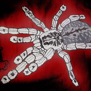 May include: A white spider with a grey body and black markings is embroidered on a red fabric background.