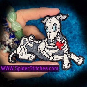 May include: Embroidered patch featuring a skeleton dog holding a red heart. The dog is white with gray accents, and the patch has a black border. The website address WWW.SpiderStitches.com is printed at the bottom of the patch.