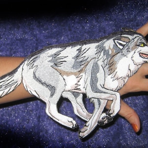Puede incluir: Un parche bordado de un lobo gris y blanco con ojos amarillos. El parche está sujeto al brazo de una persona.