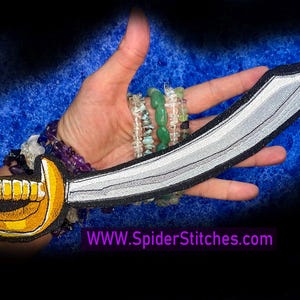 May include: A curved sword patch featuring a silver blade with black outlines and a yellow hilt with a gold guard. The patch is embroidered with detailed stitching. The text WWW.SpiderStitches.com is visible in purple at the bottom of the image.