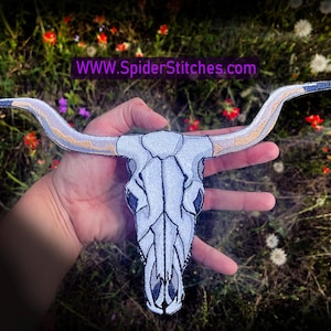 Op de afbeelding: Een witte geborduurde patch met een stieren schedel met gouden hoorns. De patch wordt in een hand gehouden tegen een achtergrond van groen gras en witte en rode bloemen. De tekst "WWW.SPIDERStitches.com" is zichtbaar bovenaan de afbeelding.