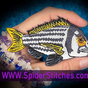 Puede incluir: Un parche bordado en blanco y negro de un pez con aletas amarillas y un ojo naranja. El pez está en una mano con un brazalete de piedras de colores. El texto "WWW.SpiderStitches.com" está en la parte inferior de la imagen.