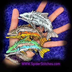 Puede incluir: Parches de peces bordados en blanco, marrón, naranja, amarillo, verde y azul. Cada parche presenta costuras detalladas y un diseño único. Los parches se muestran en una mano sobre un fondo azul. Texto: WWW.SpiderStitches.com.