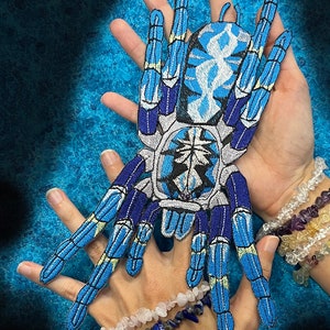 Op de afbeelding: Blauw-witte geborduurde vogelspin-patch met gele accenten. De patch wordt in de handen van een persoon gehouden. De tekst "WWW.SpiderStitches.com" is zichtbaar in de linkerbovenhoek van de afbeelding.