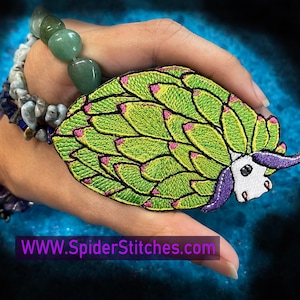 May include: A green and purple embroidered patch of a sea slug with a white body and a green shell. The patch is held in a hand with a bracelet on the wrist. The text "WWW.SpiderStitches.com" is visible in the bottom left corner of the image.