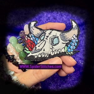 Puede incluir: Parche bordado con una calavera estilizada con cuernos, adornada con motivos florales rojos, azules y verdes. El parche tiene un borde negro y el texto "WWW.SpiderStitches.com" en un rectángulo morado.