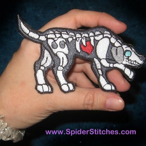 Op de afbeelding: Geborduurde patch van een hondenskelet met een rood hart. De hond is zwart en wit met een gedetailleerd botstructuurontwerp. De patch heeft een witte rand en wordt in een hand gehouden.
