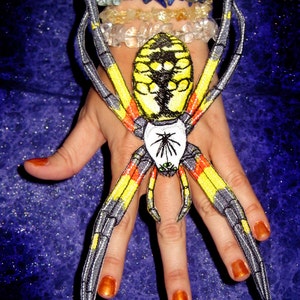 Könnte beinhalten: Eine gelbe und schwarze Spinne mit roten Streifen an den Beinen wird auf der Hand einer Person gehalten. Die Spinne ist ein Aufnäher oder ein gesticktes Design.