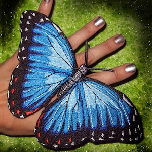 May include: A blue and black embroidered butterfly with red accents. The butterfly is on a hand with silver nail polish.