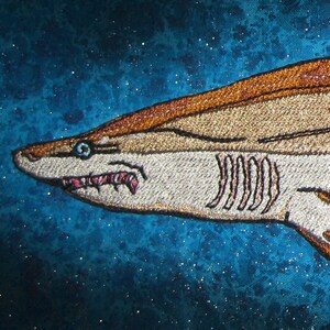 May include: Embroidered shark patch with a brown and white color scheme. The shark is facing left and has a blue eye. The background is a blue and white speckled fabric.