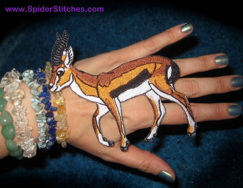 May include: Embroidered patch featuring a gazelle with brown, orange, and white details. The patch has a black outline and is set against a dark background. The website address "www.SpiderStitches.com" is visible at the top.