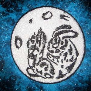 May include: A white embroidered patch featuring a black silhouette of two rabbits on a moon with three craters. The rabbits are facing each other and appear to be holding paws.
