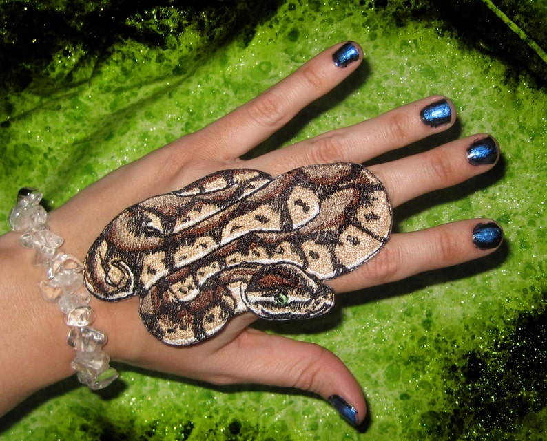 May include: A brown and tan snake patch with a green eye, sewn onto a hand with blue nail polish.