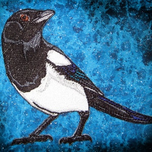 May include: A black and white magpie embroidered on a blue sparkly fabric background.