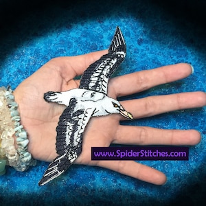 Puede incluir: Un parche de hierro bordado en blanco y negro de una gaviota en vuelo. El parche está en una mano con una pulsera de cuentas turquesa y blanca. El texto "www.SpiderStitches.com" es visible debajo del parche.
