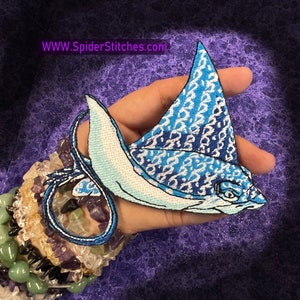 Peut inclure: Patch thermocollant brodé bleu et blanc représentant une raie manta avec un fond blanc et des écailles à motifs bleus et blancs. Le patch est sur une main avec une pile de bracelets.