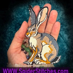 May include: Embroidered patch featuring a detailed jackrabbit design. The patch showcases a seated rabbit with a mix of gray, tan, and cream colors, accented with orange and black details. The patch has the text "WWW.SpiderStitches.com" at the bottom.