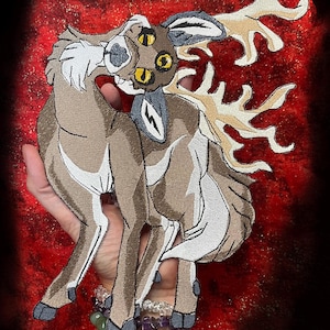 Puede incluir: Parche bordado con una criatura parecida a un ciervo con tres ojos y grandes cuernos. El parche es principalmente marrón, con detalles blancos y grises. El texto "WWW.SpiderStitches.com" es visible en la parte superior.