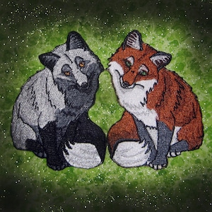 May include: Two embroidered foxes, one silver and one orange, are sitting side by side. The silver fox has black eyes and a black nose, while the orange fox has green eyes and a black nose. Both foxes have white fur on their chests and tails.