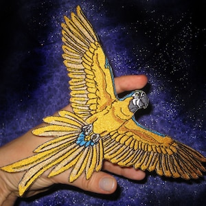 May include: A yellow and blue embroidered patch of a macaw in flight. The macaw has a yellow body, blue wings, and a black beak.