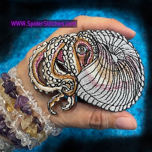 Könnte beinhalten: Gestickter Aufnäher eines Nautilus mit einem detaillierten, komplizierten Design. Der Nautilus ist weiß mit braunen und rosafarbenen Akzenten. Der Aufnäher wird in einer Hand mit einem Armband aus bunten Steinen gehalten.