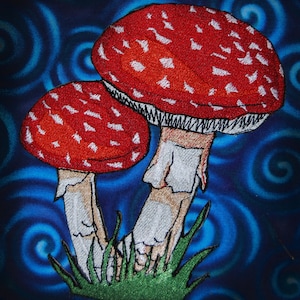 May include: Two red and white mushrooms with white stems and green grass at the base, embroidered on a blue background with swirling patterns.