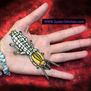 Peut inclure: Un écusson thermocollant brodé représentant une termite blanche et jaune. L'écusson est sur une main portant un bracelet turquoise. Le texte "WWW.SpiderStitches.com" est dans le coin supérieur droit de l'image.