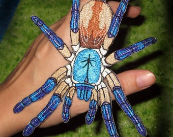 May include: Embroidered spider patch featuring a detailed design with blue, brown, and tan colors. The spider has eight legs and a detailed body. The patch is likely for clothing or accessories.